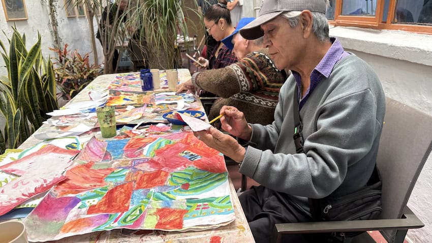 Un anciano dibujando en una hoja de papel con una obra de arte más grande sobre la mesa frente a él.